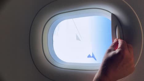 passenger hand opening aircraft window blind from which airplane wing and clouds can be seen