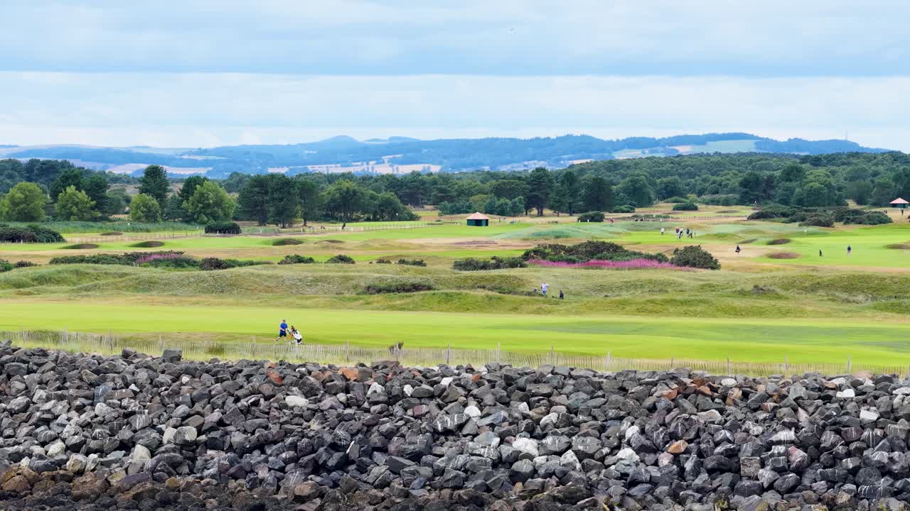 Golfers walk along green fairway, rocky foreground, overcast daylight, wide static landscape shot