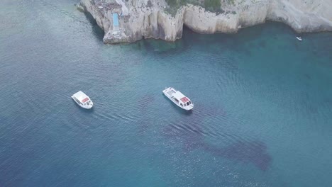 drone footage of cliffs on the coast of zakynthos greece, featuring two yachts, blue seas and several buildings on top of the cliffs