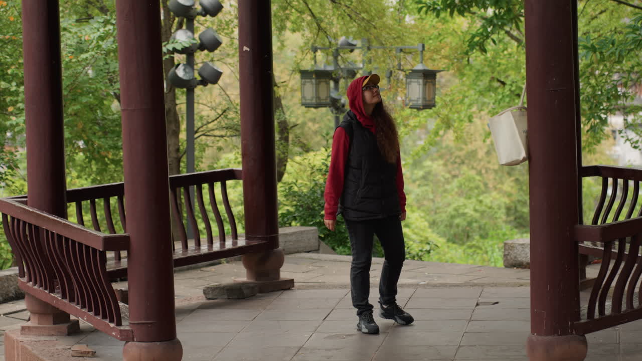 Donna che esplora un padiglione di legno nel parco con sguardo attento, giacca rossa con cappuccio, camminando tra colonne di legno, con lanterne appese e una valle lussureggiante sullo sfondo, trasmettendo un movimento casuale e una pausa osservativa.