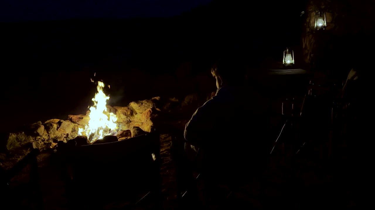 Boy sits in darkness next to roaring campfire with yellow flickering flames