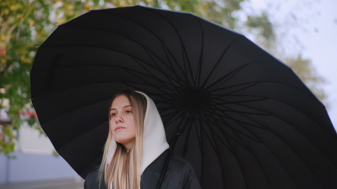 Young woman with long blonde hair wearing white hood and black coat walks outdoors under large black umbrella, looking calm and thoughtful, natural background of green trees blurred softly in daylight