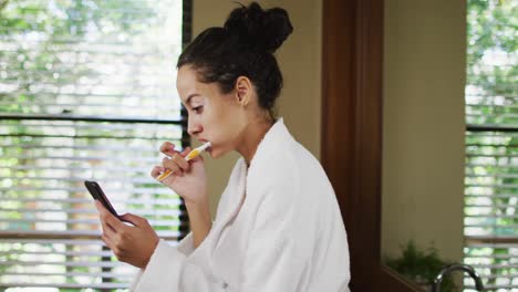 Feliz-Mujer-Birracial-En-Bata-Cepillándose-Los-Dientes-Y-Usando-Un-Teléfono-Inteligente