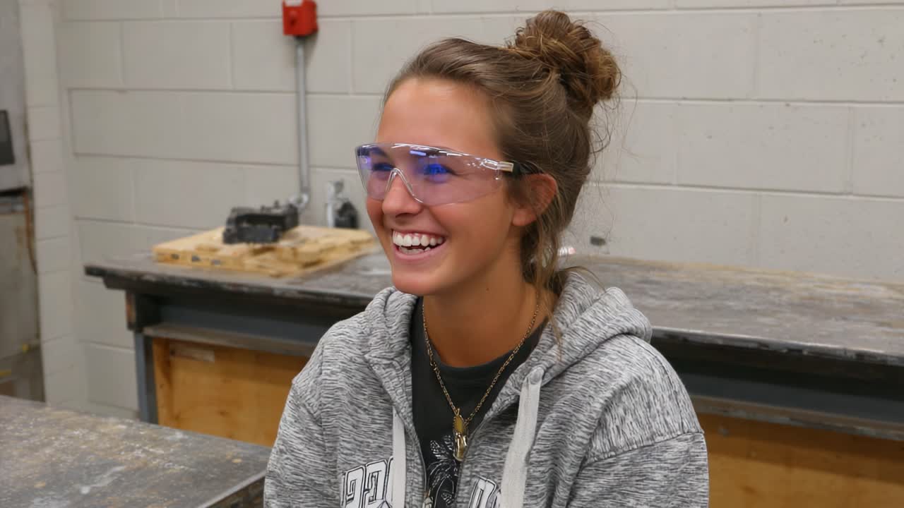 A Smiling Young Woman in Protective Eyewear Enjoys a Moment in a Workshop, Showcasing Enthusiasm and Safety in a Hands-on Environment
