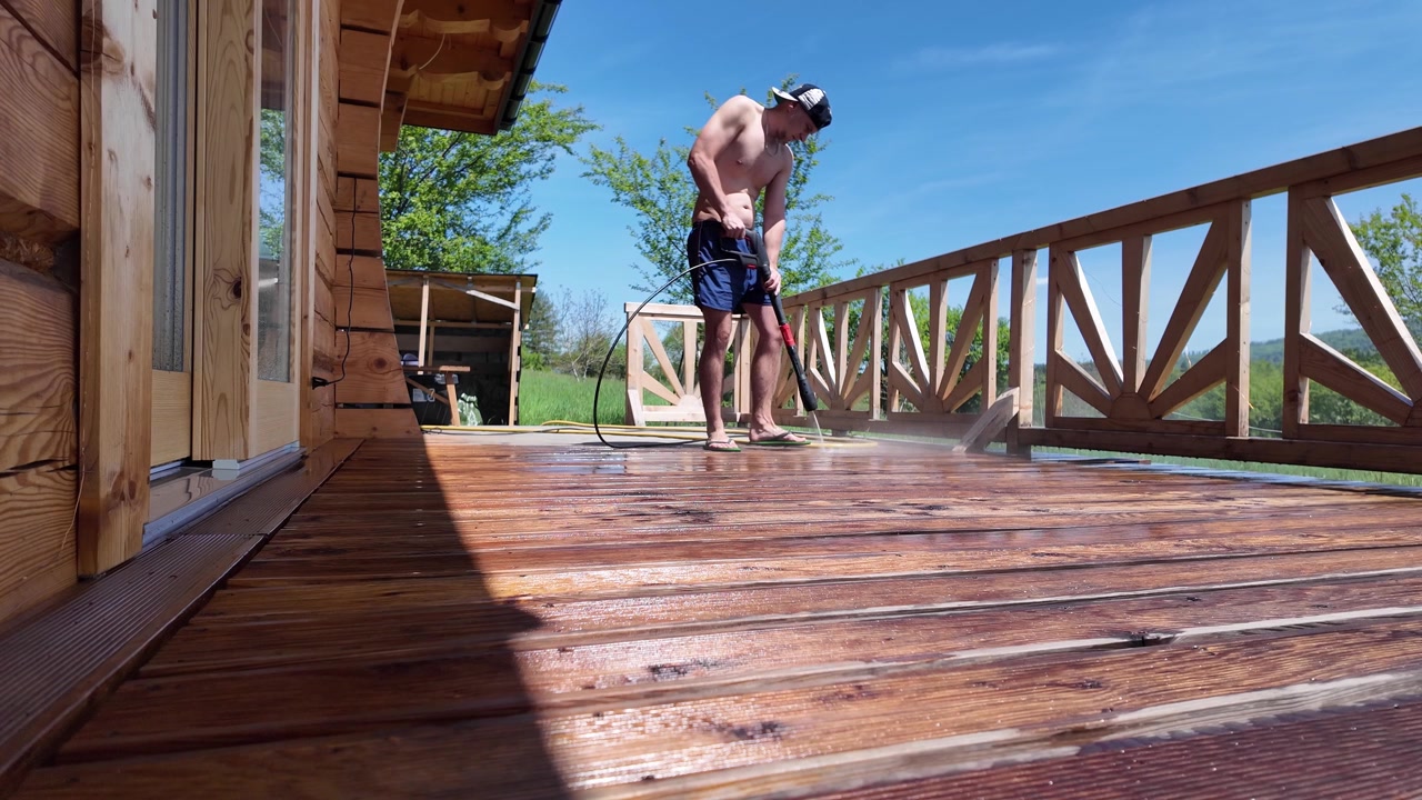 Uomo a torso nudo in pantaloncini, infradito e berretto da baseball usa un'idropulitrice per pulire una terrazza in legno presso una baita rustica in una luminosa giornata estiva di sole.