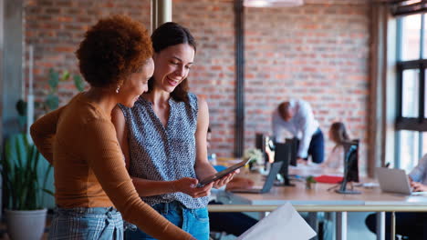 Two-Female-Business-Colleagues-With-Digital-Tablet-And-Documents-Meeting-In-Busy-Office