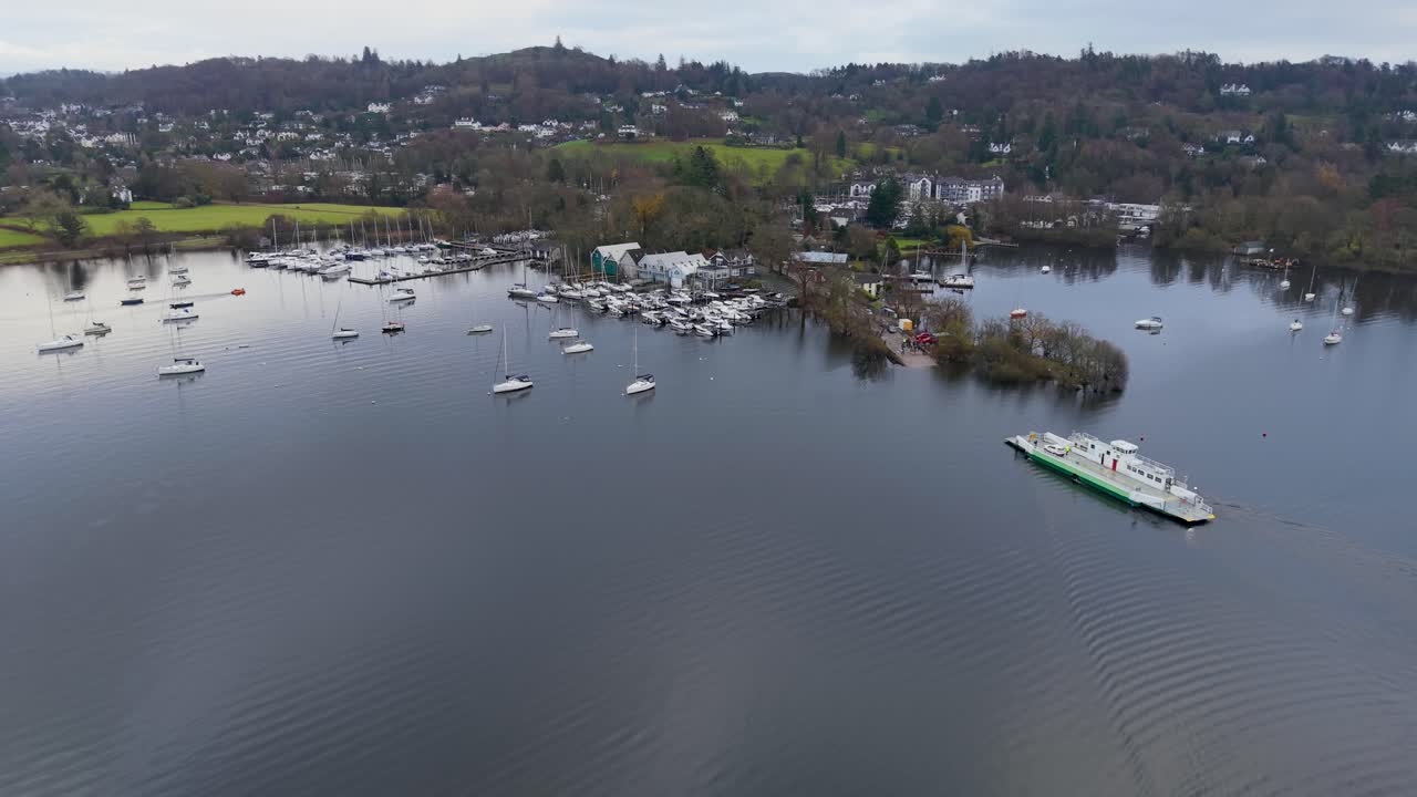 Drone turun di atas Danau Windermere, Cumbria, Inggris Raya saat feri mendekati dermaga utama Bowness. Marina berada di belakang, dengan rumah-rumah, pepohonan, dan perbukitan di kejauhan di bawah langit biru yang dipenuhi awan