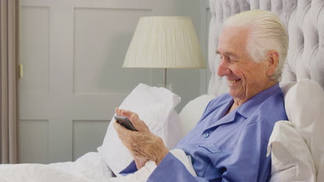 Un-Hombre-Mayor-Sonriente-En-Casa-Usando-Pijamas-En-La-Cama-Usando-Un-Teléfono-Móvil