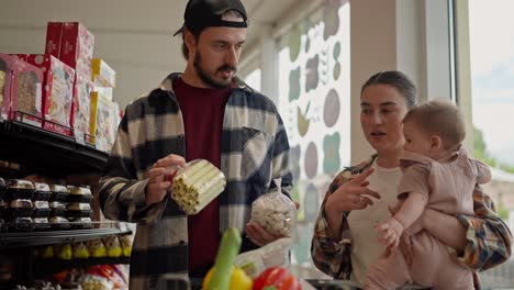 Confident-brunette-man-with-a-beard-in-a-cap-together-with-his-wife-and-small-child-choose-goods-during-their-family-shopping-in-a-supermarket