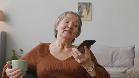 Senior-Woman-Making-Hands-Free-Phone-Call-At-Home-While-Holding-A-Coffee-Cup-Sitting-On-Sofa-At-Home