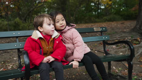 view of a little boy and a little girl smiling,closing up to pose for a photo on a black color bench in a park on a wintry evening