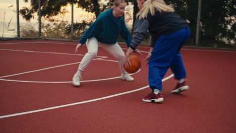 Close-up-shot-of-two-girls-playing-basketball-on-the-red-basketball-court-and-one-of-them-trying-to-throw-the-ball-into-the-basketball-hoop-in-the-morning-during-training