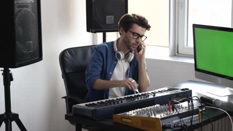 young man in sound recording studio. guy sit at keyboard and adjust sound console before record. talking on phone. green digital screen.