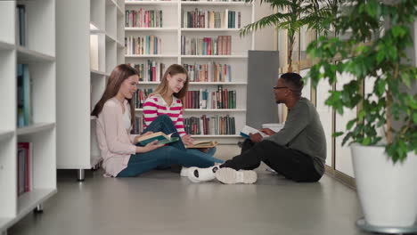amigos felices se sientan en el suelo discutiendo un proyecto de inicio en la biblioteca pública. mujeres y hombres negros leen libros juntos en la librería. grupo de estudiantes hacen la tarea juntos