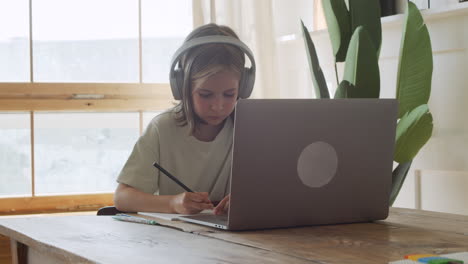 A-Pretty-And-Intelligent-Blonde-Girl-With-Headphones-Looks-At-The-Laptop-Screen-And-Does-Her-Homework-Using-A-Pencil-And-Paper