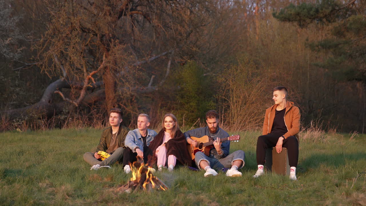Group of friends sitting near bonfire. Picnic of young people with guitar near bonfire in the evening