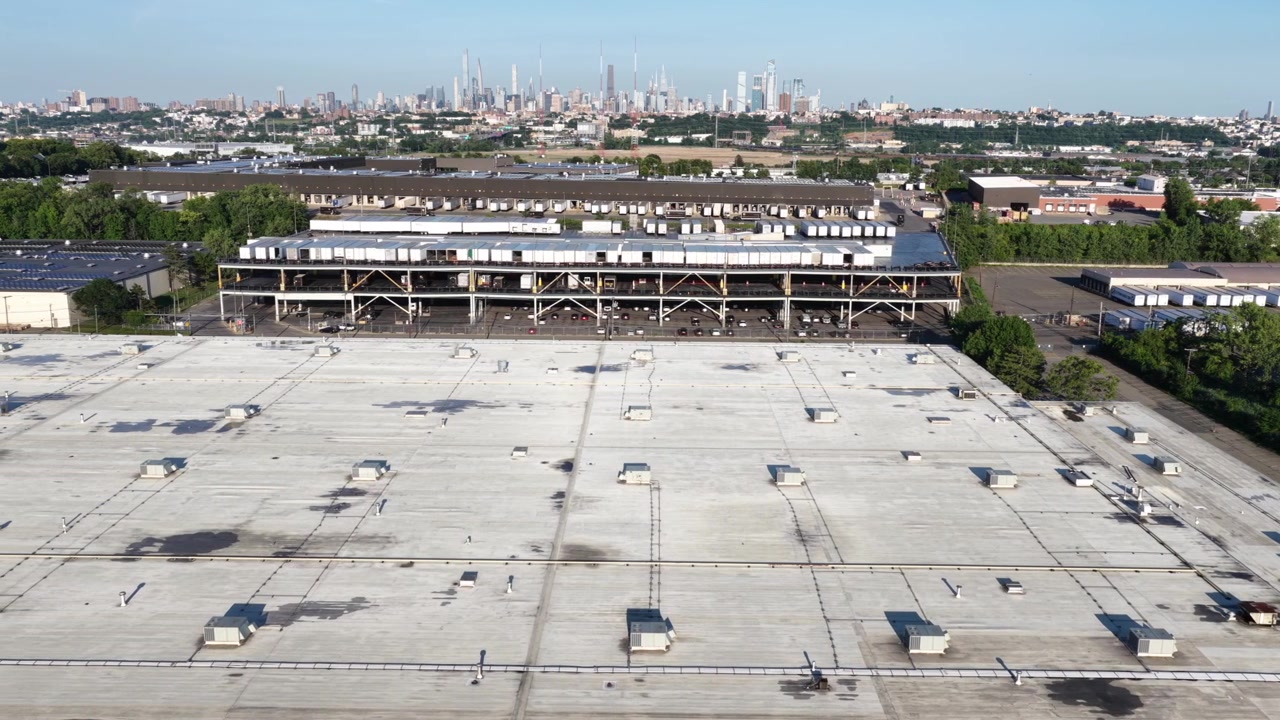 Un tejado gris plano se extiende sobre un gran almacén en Secaucus, Nueva Jersey; un toldo de muelle de carga y remolques estacionados bordean el complejo inferior, y el horizonte de Manhattan se eleva más allá. Toma aérea de dron de gran angular.