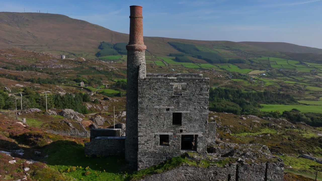 Copper Mine, Allihies, County Cork, Ireland, September 2024. Drone orbits clockwise around crumbling stone ruin with tall brick chimney on a sunny mountain slope.