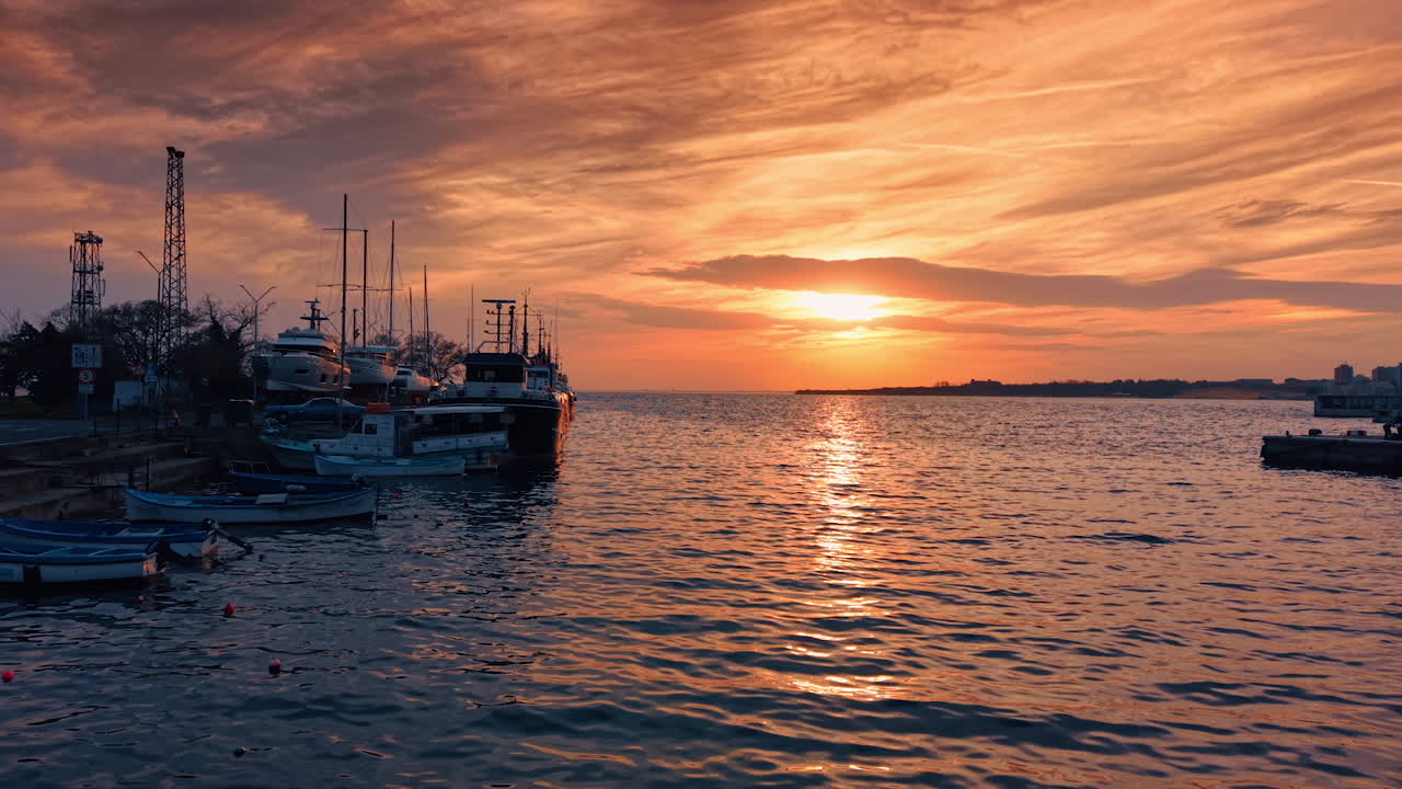 Le barche sono ormeggiate in un porticciolo vicino alla riva mentre il sole tramonta sull'acqua. Il cielo si tinge di arancione e rosa, riflettendosi sulla superficie calma.