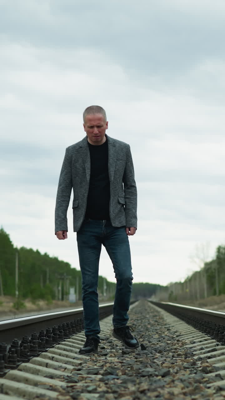 A close view of an aged man walking on a railway track, wearing a grey suit, jeans, and canvas shoes, he is looking down, surrounded by trees and electric poles, with stones on the railway