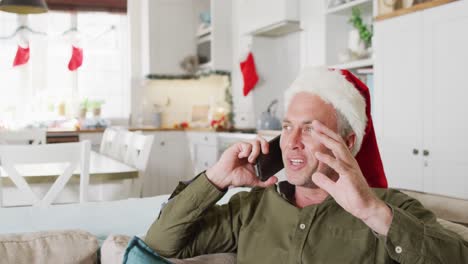 Happy-caucasian-man-wearing-santa-claus-hat,-talking-on-smartphone-in-living-room