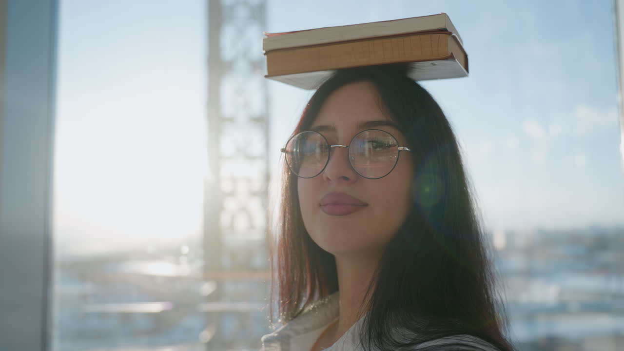 Young woman wearing glasses balances stack of books on head while standing confidently in sunlit modern glass interior, soft light casting warm glow as she gazes calmly ahead with subtle smile