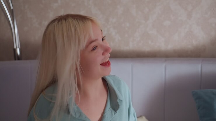 Una ragazza bionda vestita di verde balla gioiosamente seduta sul letto, esprimendo felicità e un umore spensierato durante un accogliente momento al chiuso.