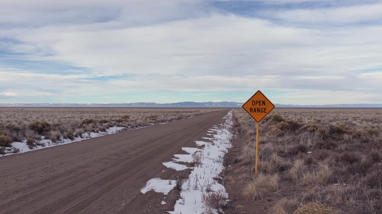 Una ripresa aerea ascendente inizia con un cartello giallo di Open Range mentre un SUV blu entra nell'inquadratura e percorre una remota strada sterrata. Questo vasto paesaggio desertico è ideale per spot pubblicitari di robusti autoveicoli.