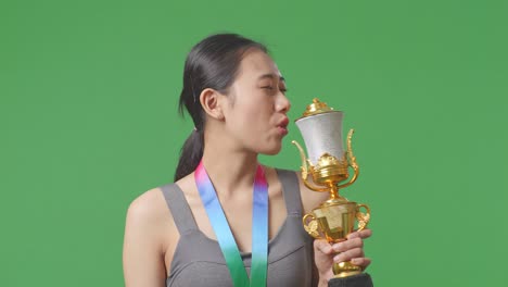 close up of asian woman with a gold medal kissing a gold trophy being proud winning as the first winner on green screen background in the studio