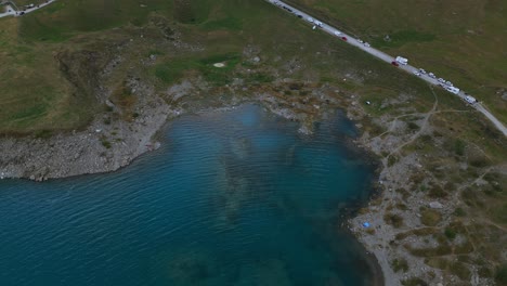 Aerial-view-of-a-turquoise-lake-with-a-road-nearby-with-cars-parked-on-the-side-of-the-road