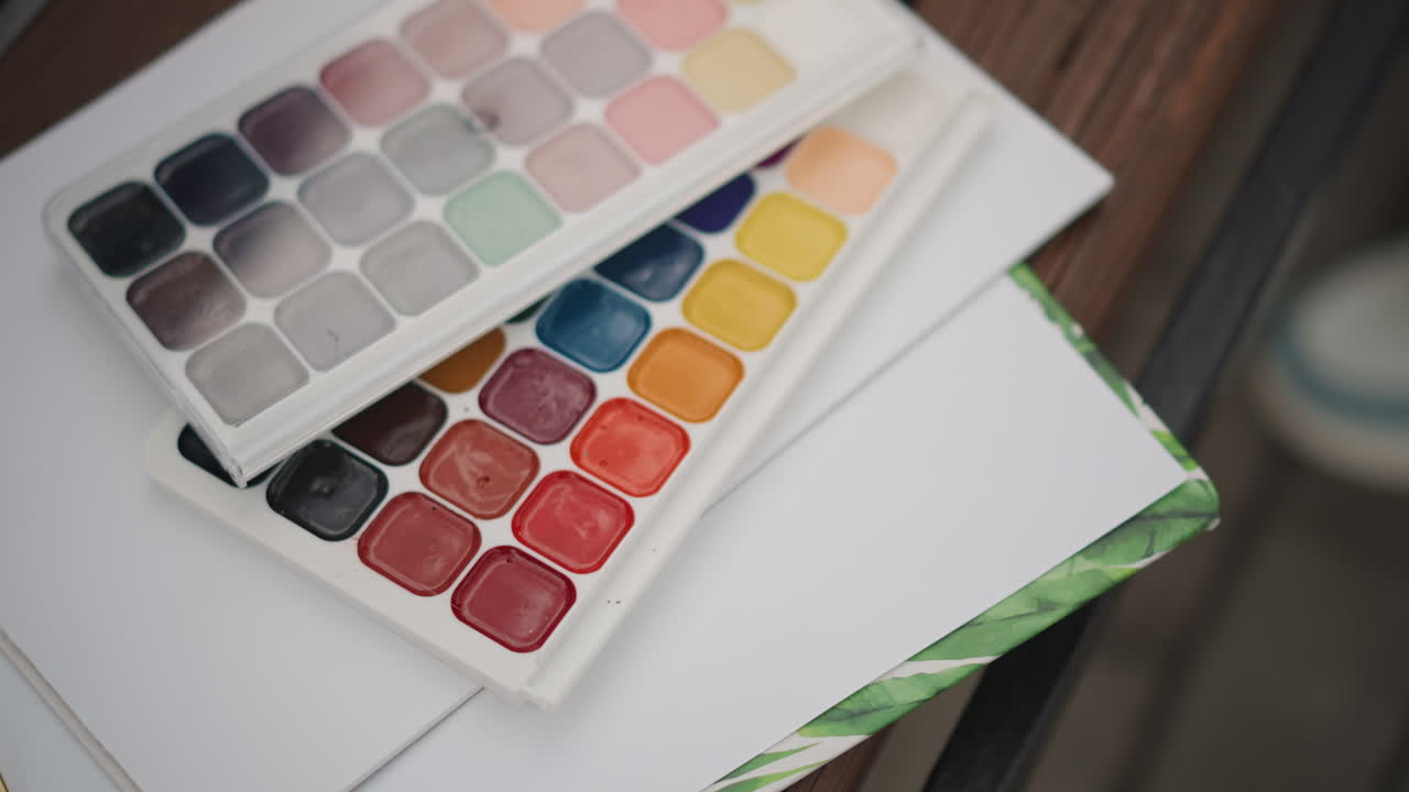 Paleta de acuarelas y cuaderno de bocetos sobre una mesa de madera, con pastillas de pigmento vibrantes, borde de papel pulcro, luz natural suave, una toma cenital de cerca que captura el espacio de trabajo del artista y sus herramientas creativas.