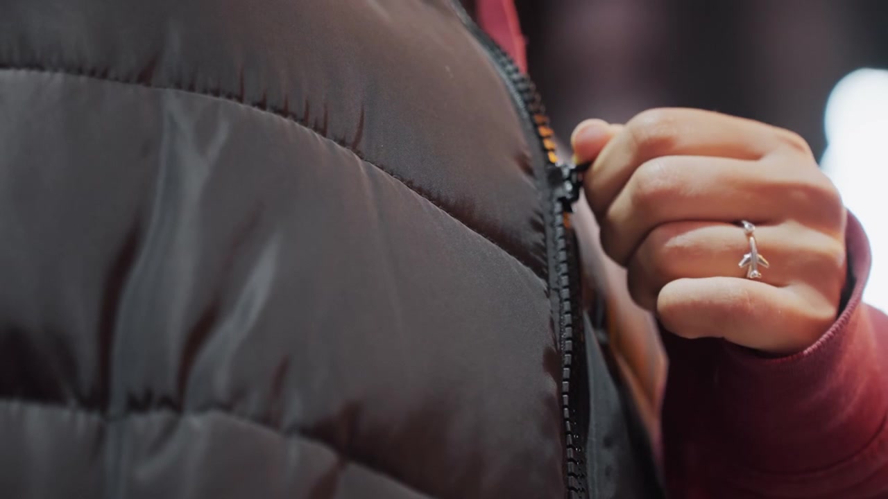 Night jacket closeup of zipper, hand with silver ring adjusting puffer near neon bokeh, subtle motion and cool evening mood, commuter pause on sidewalk showcasing textured fabric and metal teeth