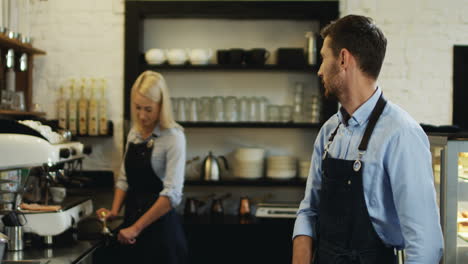 Portrait-Of-The-Young-Handsome-Waiter-Standing-In-Front-Of-The-Camera-And-Watching-Behind-At-The-Young-Blond-Waitress-Making-Coffee-On-The-Special-Machine,-Then-Smiling-To-The-Camera