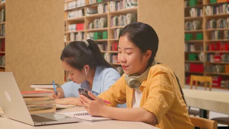 asian woman student with headphones enjoys using smartphone while sitting with her classmate studying on a table with laptop in the library