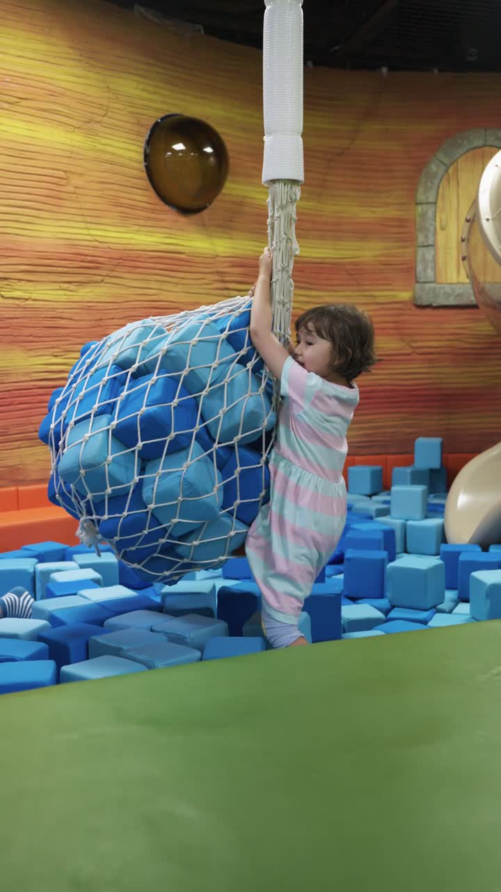 Side view of a concentrated young girl holding onto a suspended blue foam block sack above a pit of blue blocks