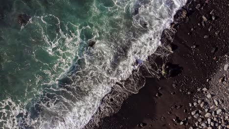 Top-down-Revealing-shot-of-mesmrising-turquoise-ocean-waves-reaching-dark-sand-shore-covered-with-small-stones-and-pebbles-closeup-to-wide-from-above