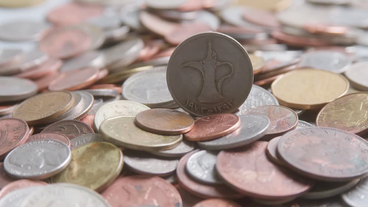Assorted international coins from various countries and denominations, different sizes, values, colours. United Arab Emirates dirham coin central emphasis. Slow camera movement, extreme close up