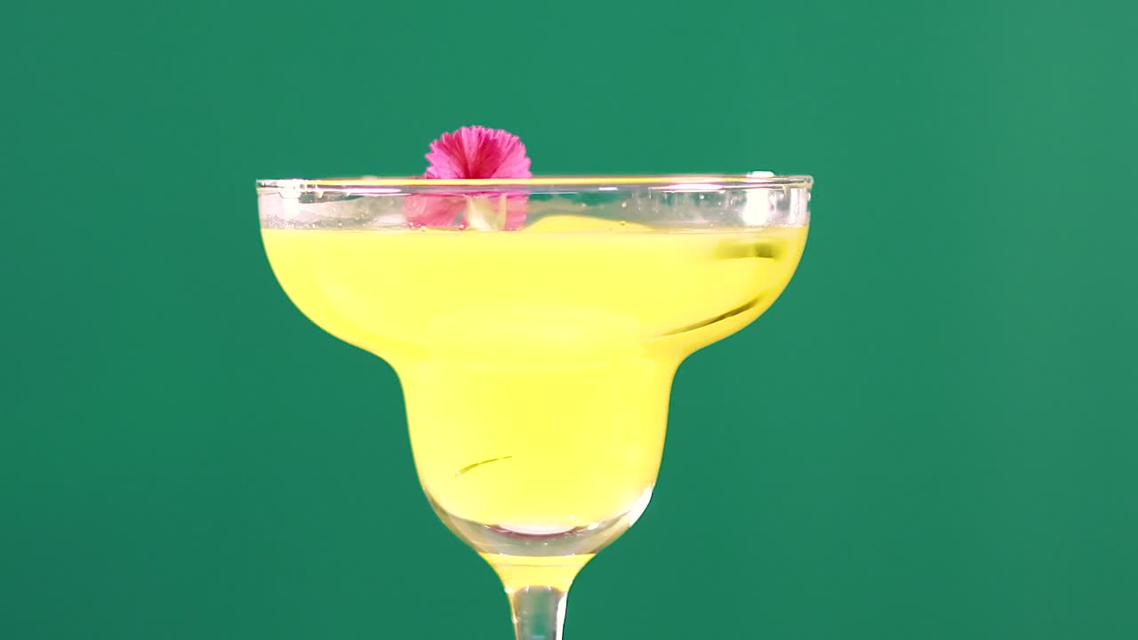 Um coquetel amarelo vibrante em uma taça de margarita com uma decoração floral rosa sobre um fundo verde