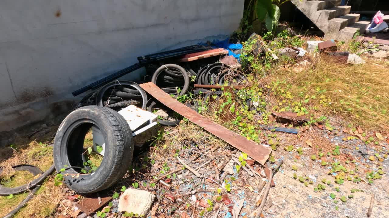 Camera pans across sunlit, littered outdoor area with tyres, debris, and overgrown vegetation