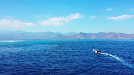 Boat-sailing-across-deep-blue-sea-toward-shore-of-mountain-tropical-island-on-a-bright-sky-with-white-static-clouds-background,-Bali