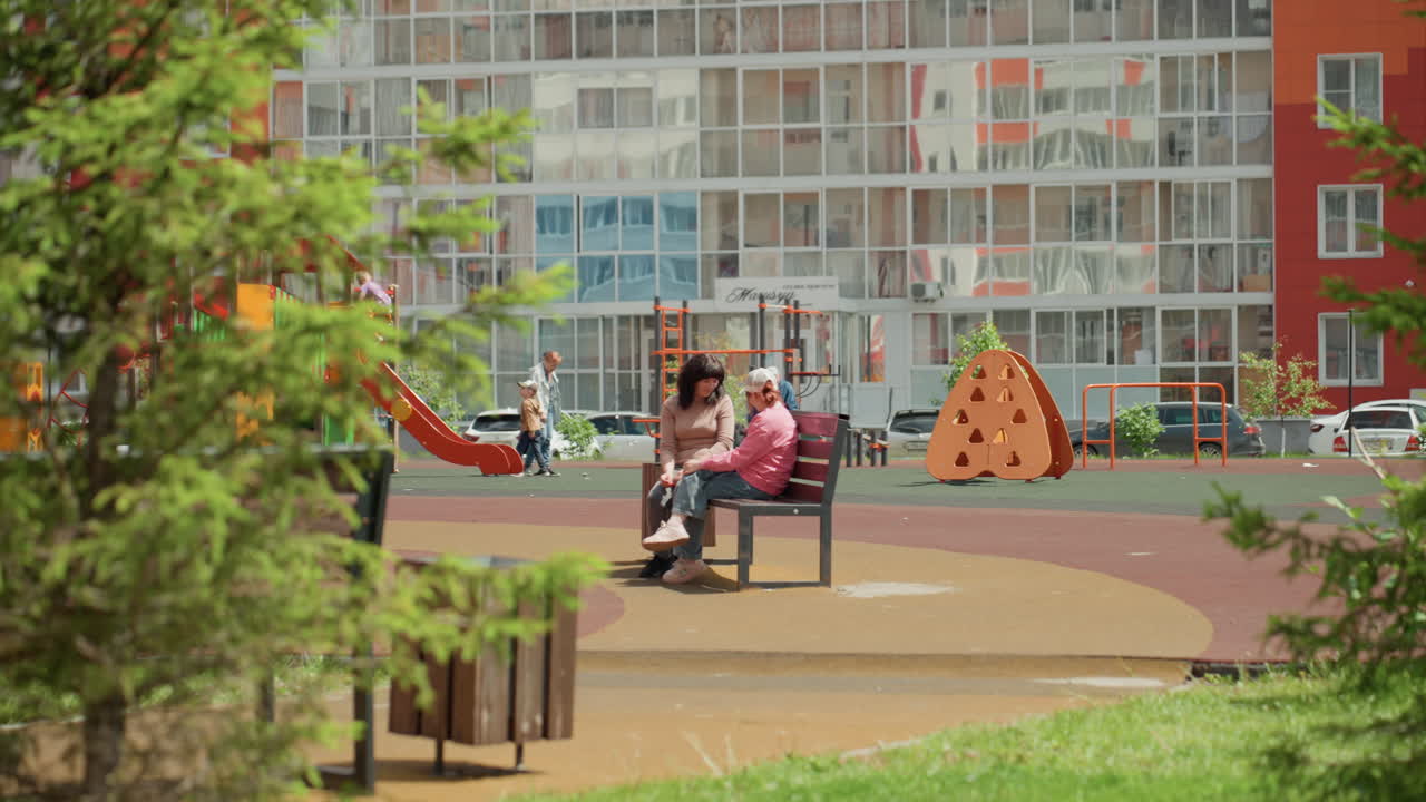 Caucasian Friends Share Quiet Playground Moment On Bench Under Trees, Apartment Facades And Colorful Play Equipment Visible, Relaxed Leisure And Soft Green Framing, Gentle Conversation And Shared