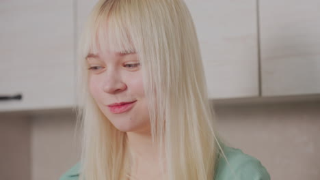 close up of young blonde girl with straight hair chewing bread with neutral expression in modern kitchen setting, cabinets behind her in soft background light