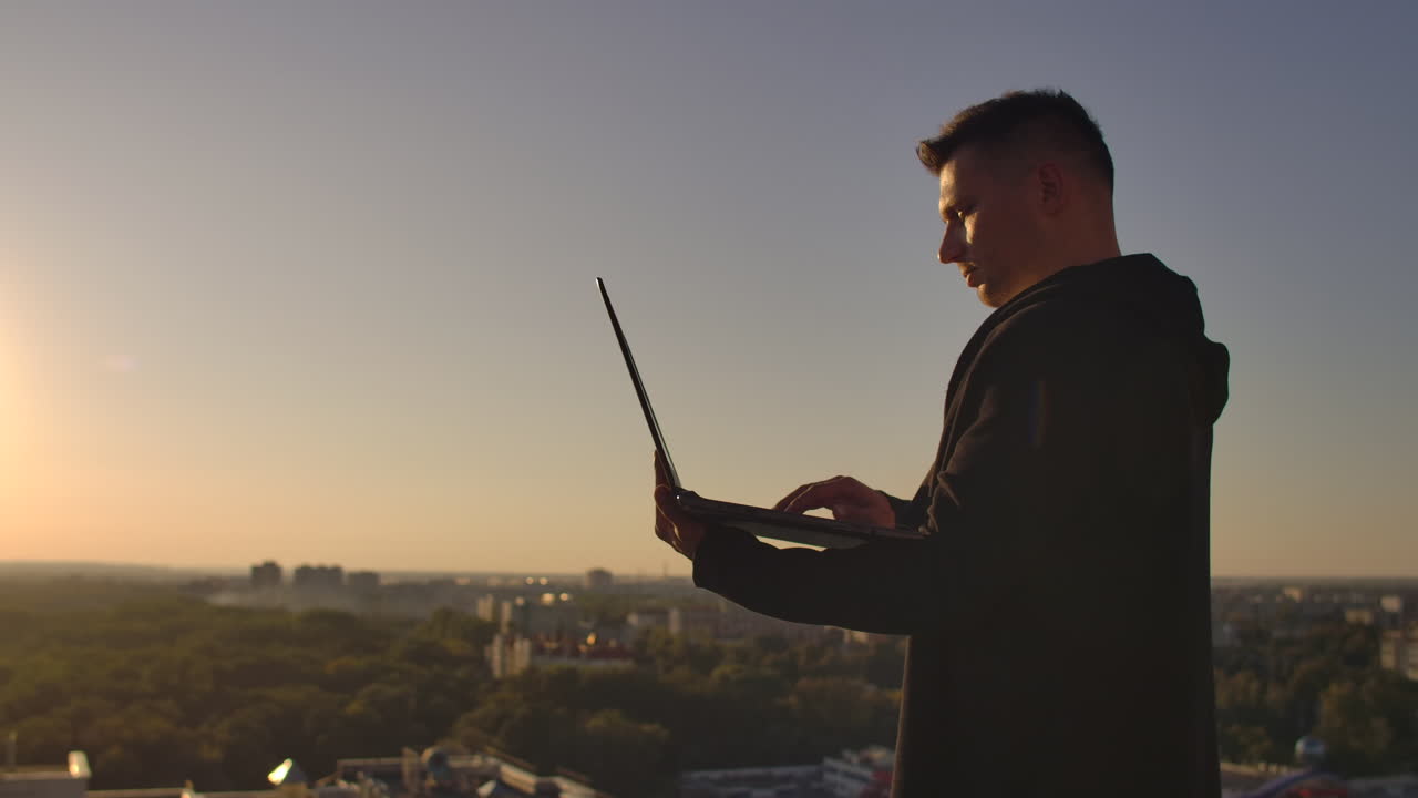 Programmer a hacker is on the roof with a laptop at sunset says error code on the keyboard and looking at the city view.