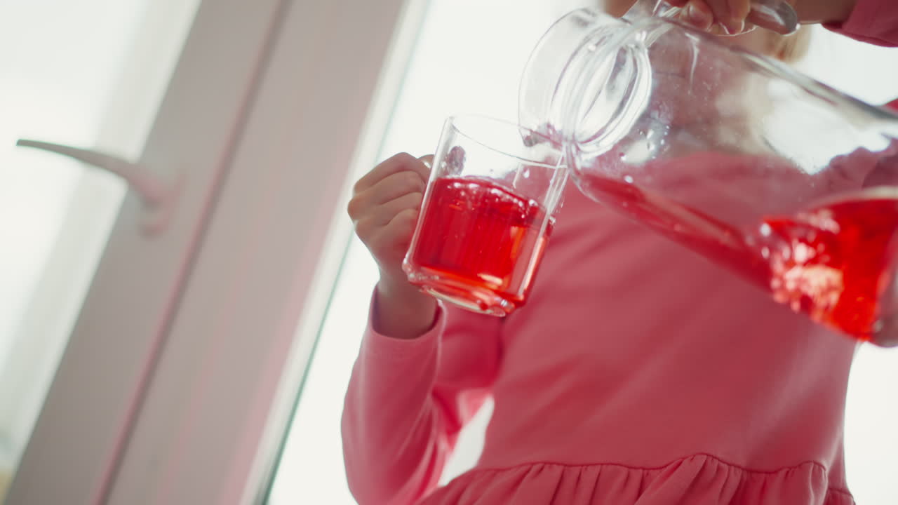 Close up of youngster in pink gown pouring vibrant red juice from glass jar into glass then sipping gently from glass indoors by sunlit window, hands gripping pitcher and cup with careful motion
