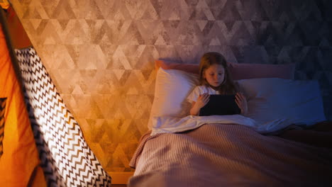 Blonde-little-girl-uses-tablet-in-bed-near-wigwam-at-night