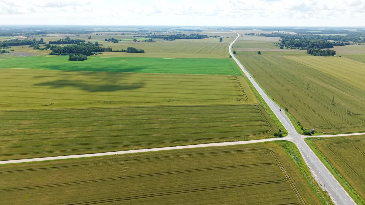 Aerial view of wide grain fields with visible tractor lines and a long road cutting through the countryside landscape