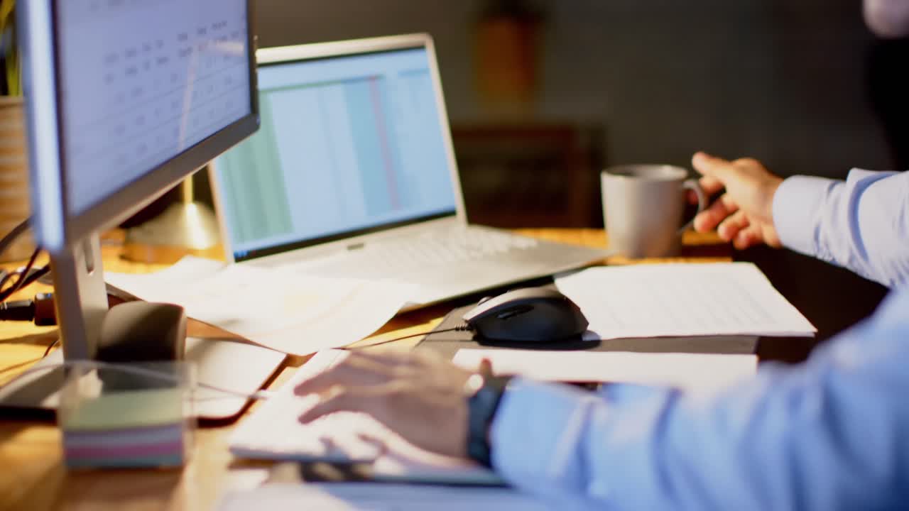 Tired biracial businessman at desk using computers, drinking coffee in office at night, slow motion