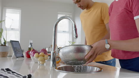 Pareja-Diversa-Preparando-Y-Lavando-Verduras-Frescas-En-La-Cocina-Con-Tableta,-Cámara-Lenta