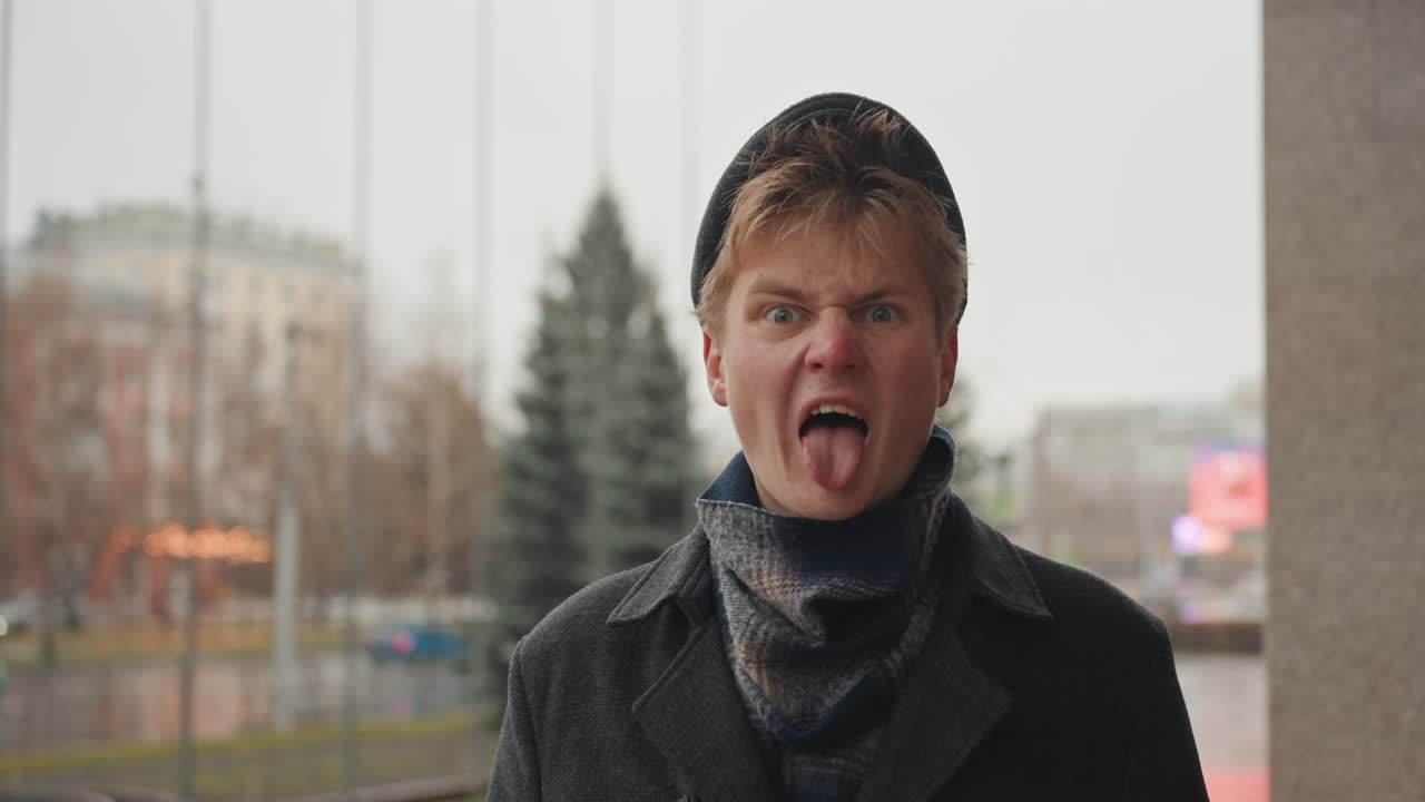 Retrato urbano juguetón de un creador masculino blanco reaccionando con expresiones animadas, gorro y bufanda, ligera nevada, luces de la ciudad borrosas, primer plano espontáneo para contenido social, con un ambiente de cineasta aspirante.