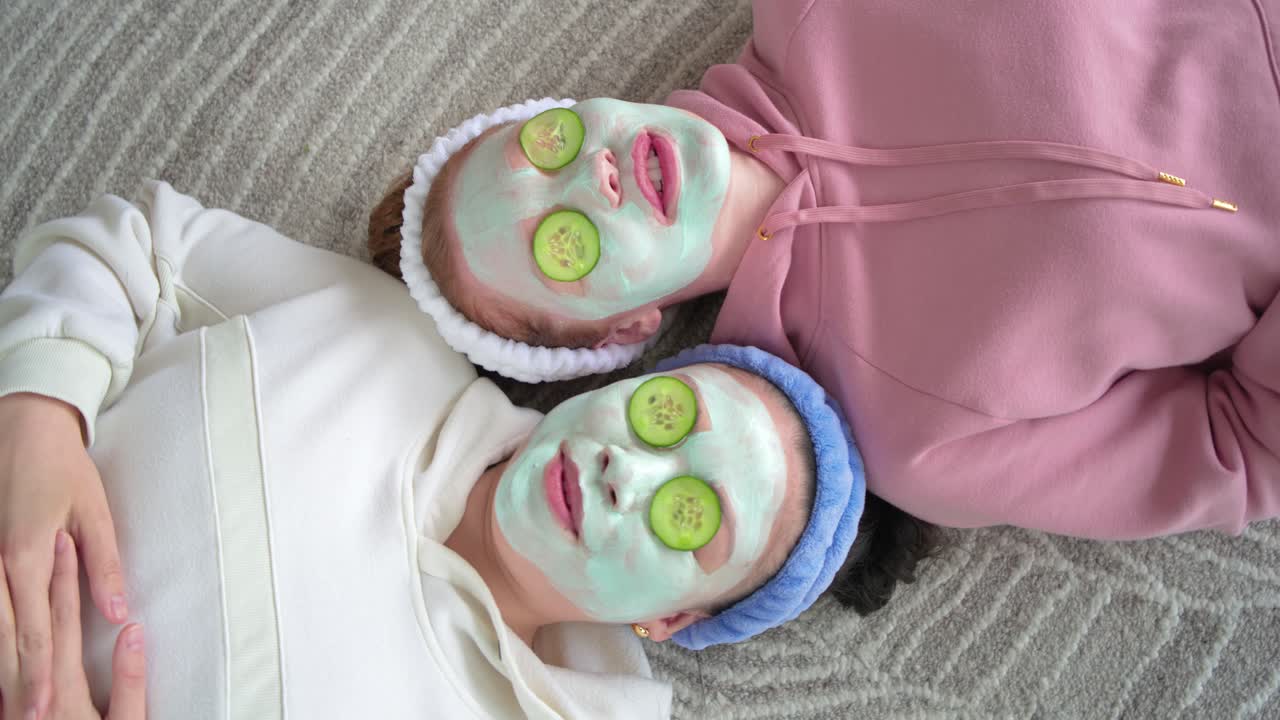Female friends lying on rug at home, one reaching adjusting cucumbers, wearing masks and laughing
